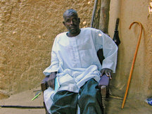 Ein afrikanischer Mann in weißem Hemd und türkiser Hose sitzt auf einem Sessel, neigt sich zur Seite und blickt in die Kamera.