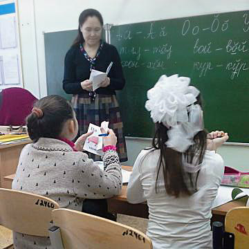 Eine Lehrerin steht vor einer beschriebenen Tafel, vor ihr sitzen zwei Schulmädchen mit heller Kleidung und dunklen Haaren.