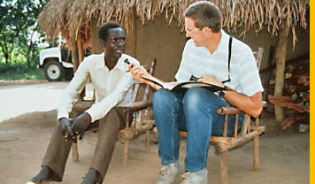 Hütte mit Strohdach, dahinter ein Auto. Davor sitzt ein afrikanischer Mann. Ein hellhäutiger Mann hält ihm ein Mikrofon hin.