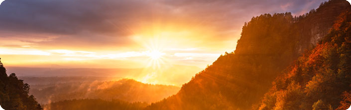 Foto: Unter graublauen Wolken strahlt die Sonne hell in Gelb und Orange über eine baumbewachsene Berglandschaft.